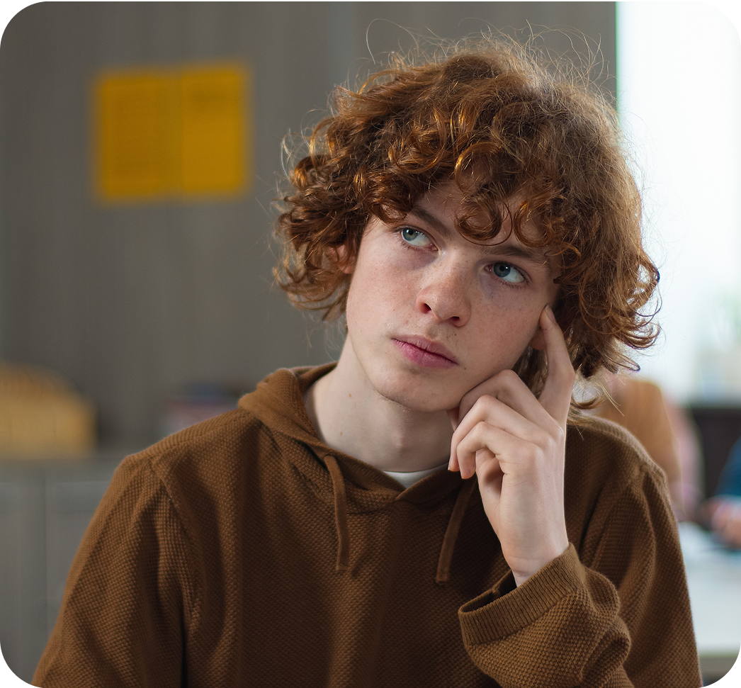 Un ragazzo adolescente con capelli ricci rossi, seduto in classe, guarda in alto pensieroso con la mano sul viso.