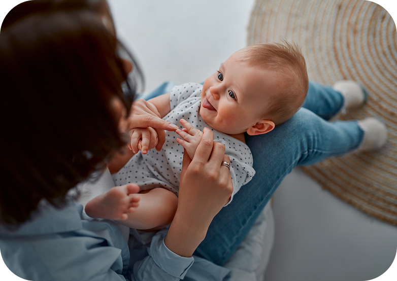 Un neonato in body bianco sorride guardando la madre che lo tiene in braccio e gli accarezza le mani, seduta su un tappeto.