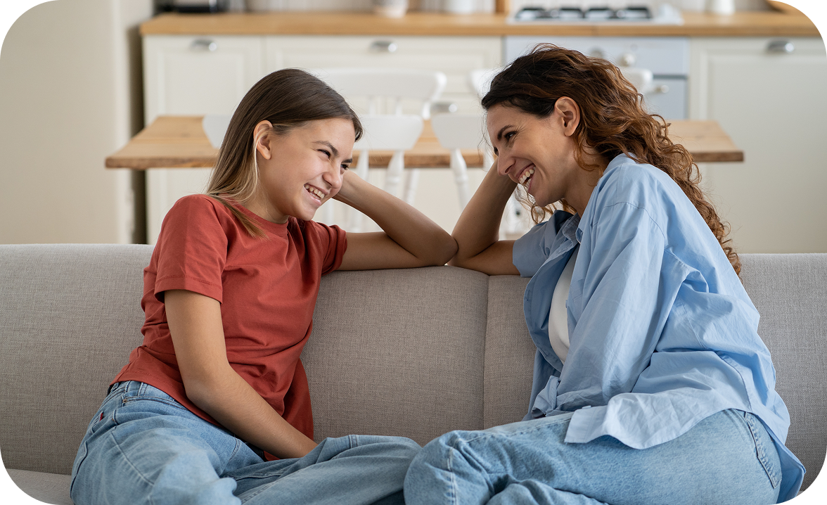 Una madre e sua figlia adolescente ridono insieme sedute sul divano di casa, in un momento di intimità e confidenza.